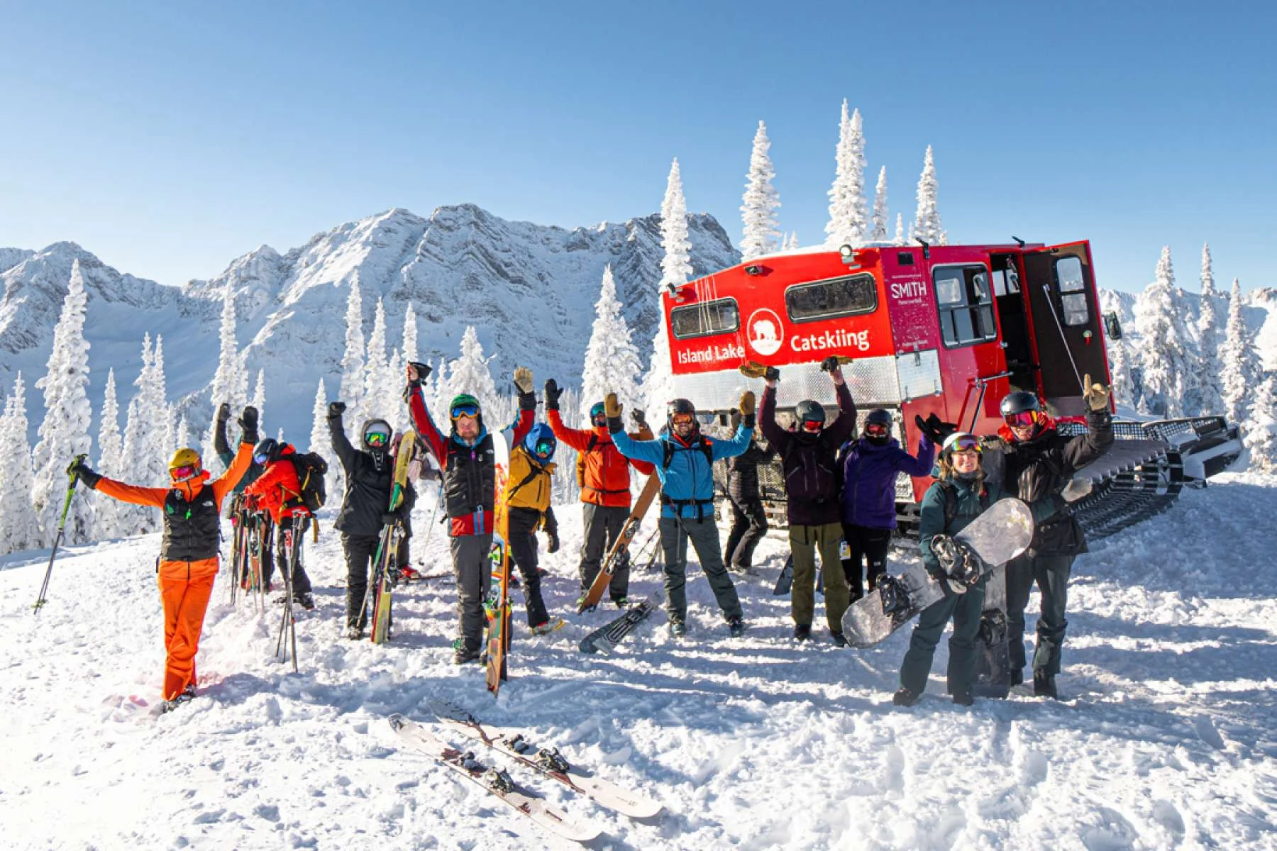 Skiers and snowboarders smiling beside a red Island Lake snowcat under snowy peaks