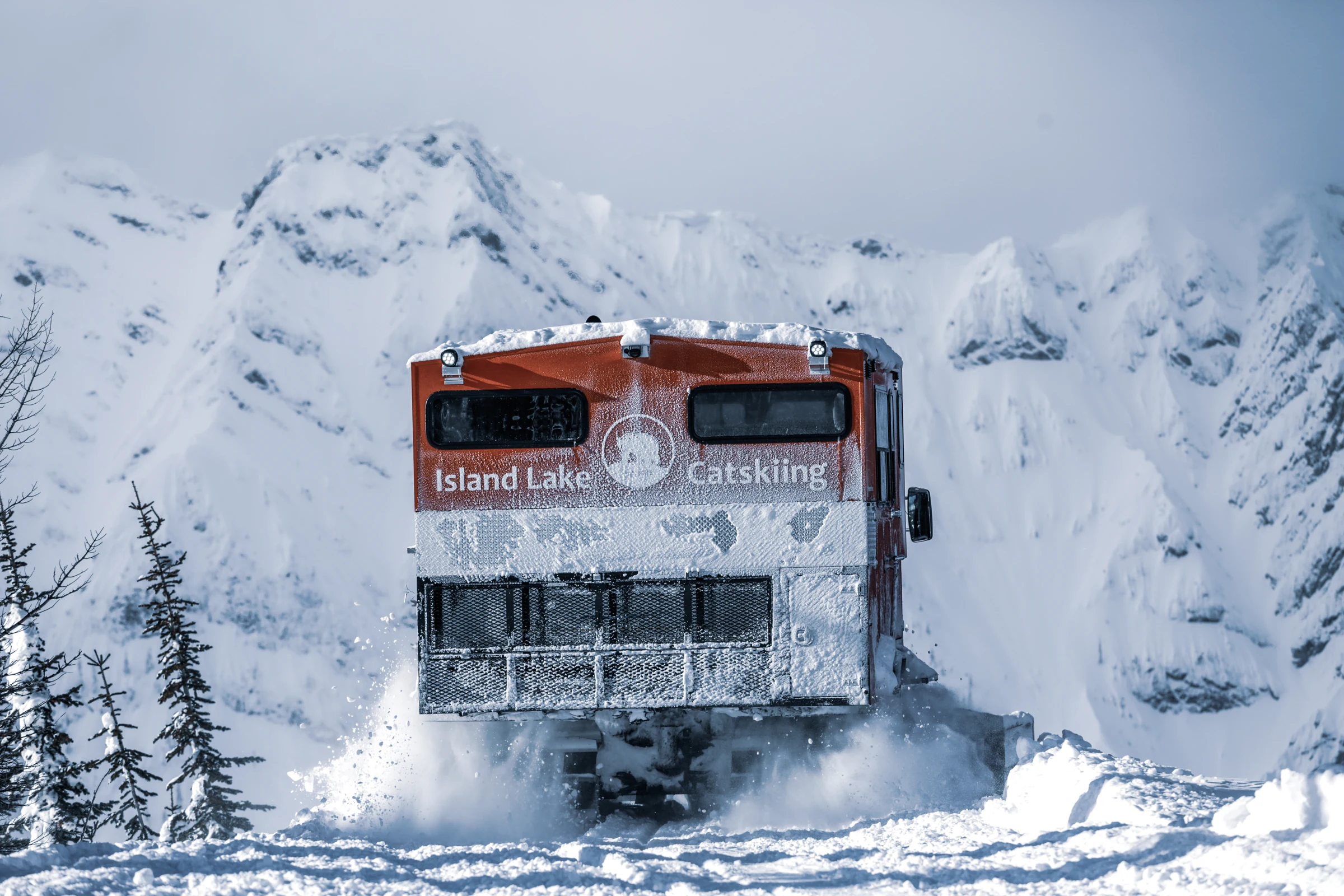 A red Island Lake snowcat climbing through snow beneath the Lizard Range