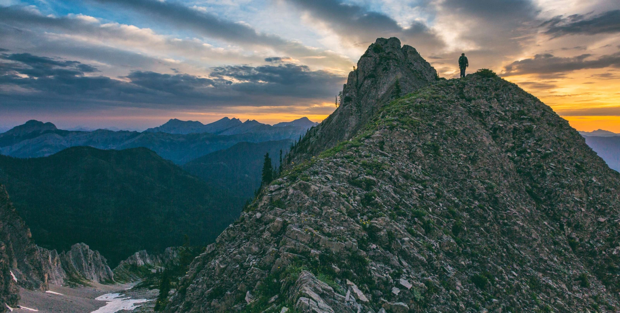 A lone hiker on a rocky alpine ridge beneath a glowing summer sunset